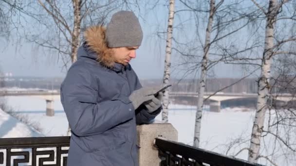 Mann in blauer Daunenjacke mit Fellkapuze nutzt Handy zum Web und plaudert im Winterpark. — Stockvideo