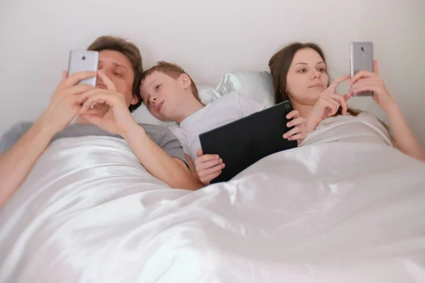 La familia está utilizando teléfonos móviles y tabletas que yacen en la cama por la mañana . — Foto de Stock