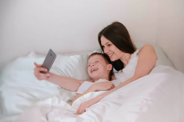 Mamá e hijo toman selfie en su teléfono móvil. Mañana en la cama . — Foto de Stock