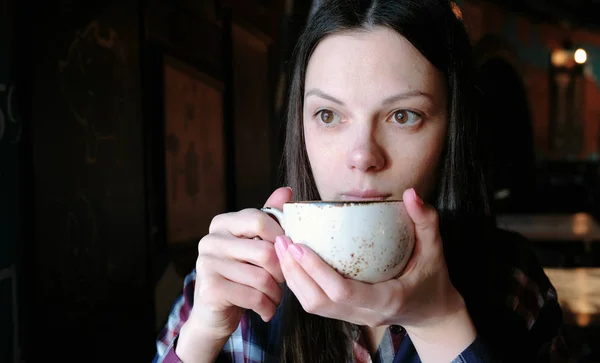 Брюнетка в клетчатой рубашке пьет чай из большой чашки. Уставшая женщина пьет чай. . — стоковое фото