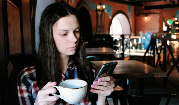 Brünette Frau im karierten Hemd trinkt Tee aus einer großen Tasse und schaut aufs Telefon. — Stockfoto