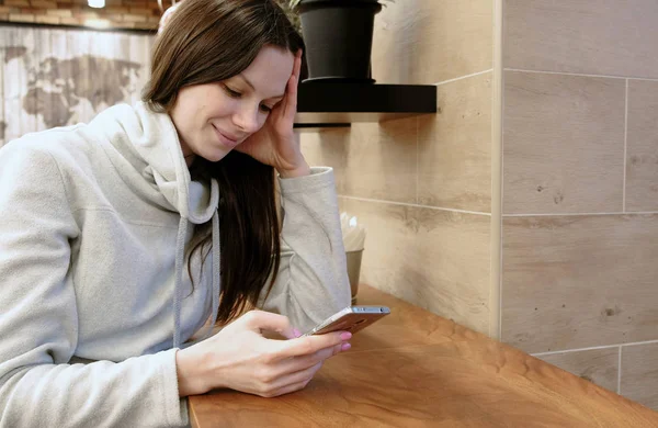 Sonriente joven morena con una camisa brillante masajeando en su celular sentada en la cafetería . — Foto de Stock