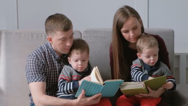 家族のお母さん、お父さんと 2 つの双子の兄弟は、ソファに座って本を読みます。家族の読書の時間. — ストック動画