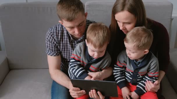 Familia con tableta. Mamá, papá y dos hijos gemelos mirando la tableta sentados en el sofá . — Vídeos de Stock