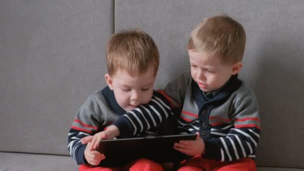 Zwei Zwillingsbrüder nehmen einander auf dem Sofa sitzend Tabletten weg. Kinder spielen Spiele auf dem Tablet. — Stockvideo