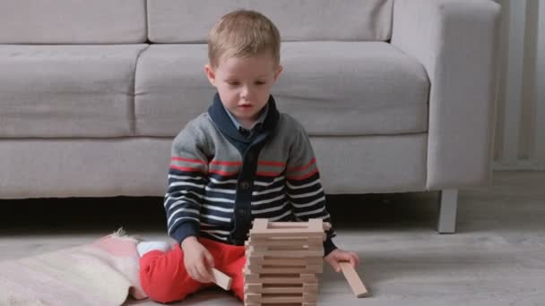 Αγόρι χτίζει ένα πύργο από ξύλινα μπλοκ κάθεται στο πάτωμα από τον καναπέ. — Αρχείο Βίντεο