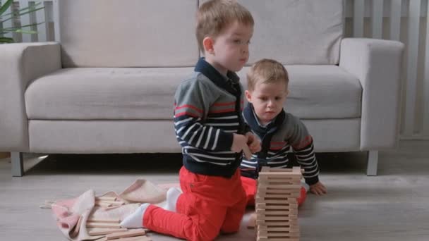 Dvojčata kluky bratři stavební věž z dřevěných kostek, sedí na pohovce v pokoji na podlaze. — Stock video