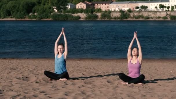 Dos mujeres estirando yoga en la playa junto al río en la ciudad. Hermosa vista de la ciudad. Postura de Namaste . — Vídeos de Stock