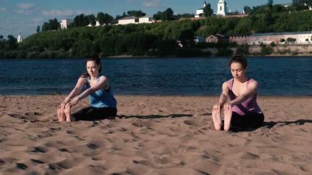 Две женщины занимаются йогой на пляже у реки в городе. Прекрасный вид на город. Пасхиманасана, поза Сасана . — стоковое видео