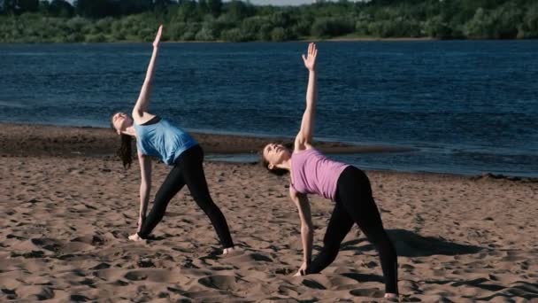 Dos mujeres haciendo yoga en la playa de arena junto al río en la ciudad. Hermosa vista de la ciudad al amanecer. Trikonasans Utiha pose . — Vídeo de stock