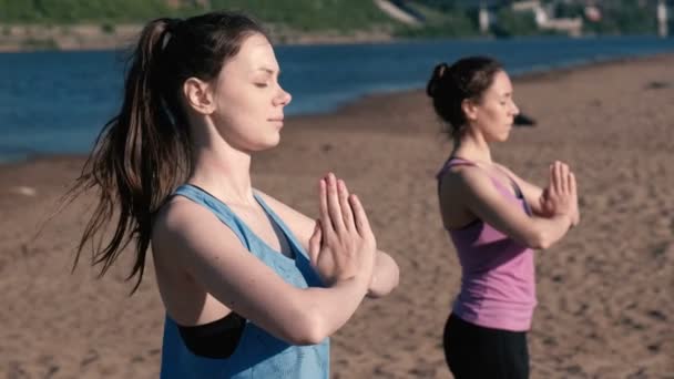 Dos mujeres estirándose yoga de pie en la playa junto al río en la ciudad. Hermosa vista de la ciudad. Postura de Namaste . — Vídeo de stock