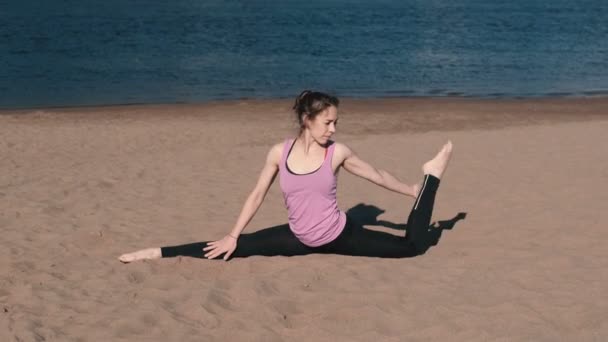 Mujer haciendo estiramiento sentado en cordel en la playa de arena al atardecer . — Vídeos de Stock