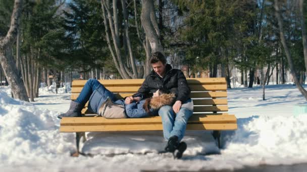 Un hombre y una mujer descansan juntos en un banco en el parque de invierno de la ciudad. Día de invierno soleado . — Vídeos de Stock