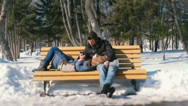 Un hombre y una mujer descansan juntos en un banco en el parque de invierno de la ciudad. Día de invierno soleado. Mira algo en el teléfono móvil . — Vídeos de Stock