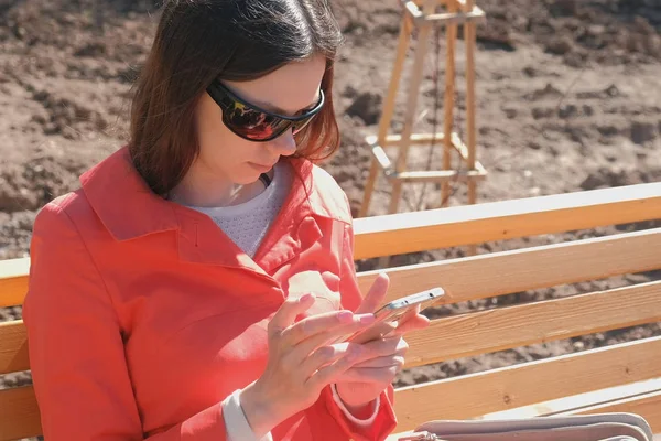 Молодая брюнетка в солнечных очках пишет и отправляет сообщение по телефону, сидя в парке на скамейке запасных. . — стоковое фото