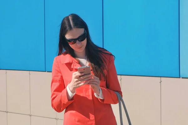 Mujer joven morena en gafas de sol en abrigo rojo espera a alguien y comprueba su teléfono, mensajes de texto . — Foto de Stock