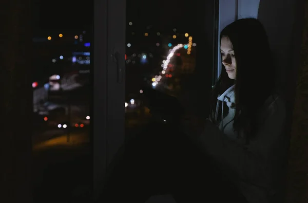 Женщина просматривает паутину на планшетном компьютере, сидя на подоконнике в темную ночь. Дорожный фон . — стоковое фото