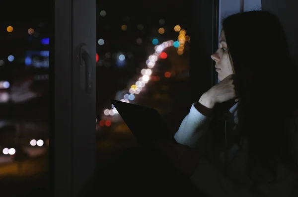 Mujer sentada en el alféizar de la ventana en la noche oscura con los ojos cerrados. Uso de tableta para web . — Foto de Stock
