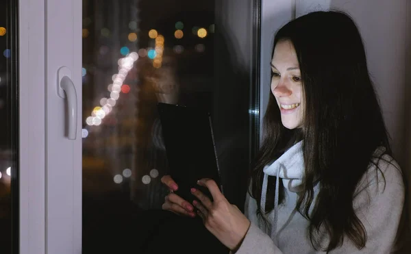 Frau schaut Film auf Tablet-Computer und lacht. in der dunklen Nacht auf der Fensterbank sitzen — Stockfoto