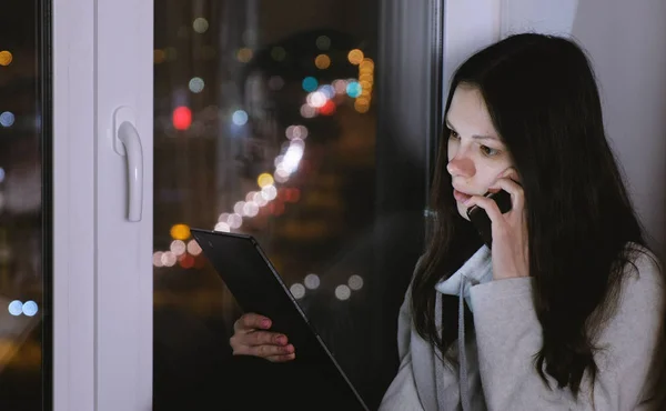 Mulher falando no celular e olhando para tablet. Sentado no peitoril da janela no escuro à noite . — Fotografia de Stock