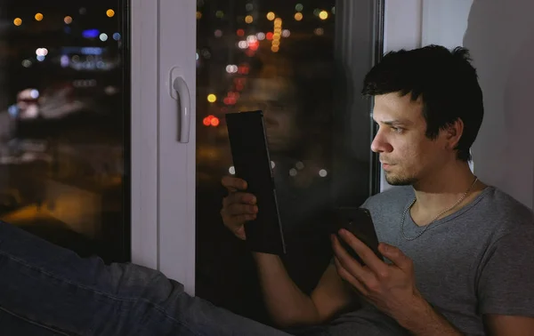 El hombre está mirando a la tableta y el teléfono móvil. Sentado en el alféizar de la ventana en la oscuridad . —  Fotos de Stock