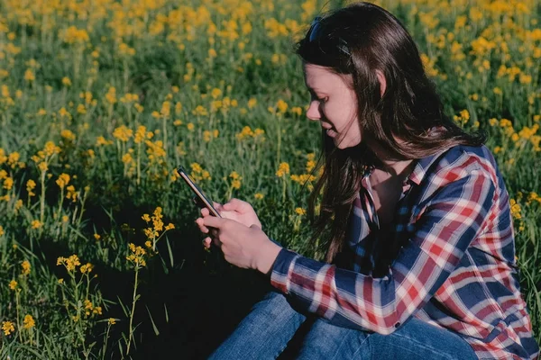 Mulher olhando para o telefone móvel sentado no parque na grama entre flores amarelas . — Fotografia de Stock