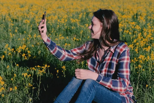 Mujer hace una selfie en el teléfono sentado en la hierba entre las flores amarillas . — Foto de Stock
