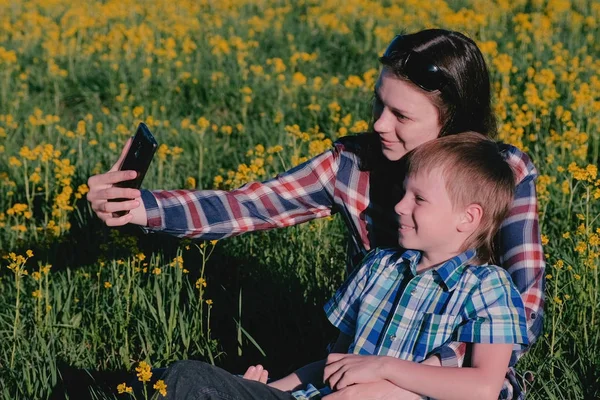Mamá e hijo hacen selfie en el teléfono sentado en la hierba entre las flores amarillas . — Foto de Stock