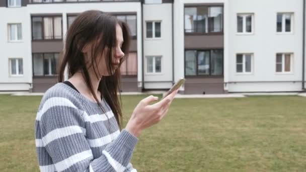 Mulher morena bonita lendo uma mensagem no celular andando perto de casa . — Vídeo de Stock
