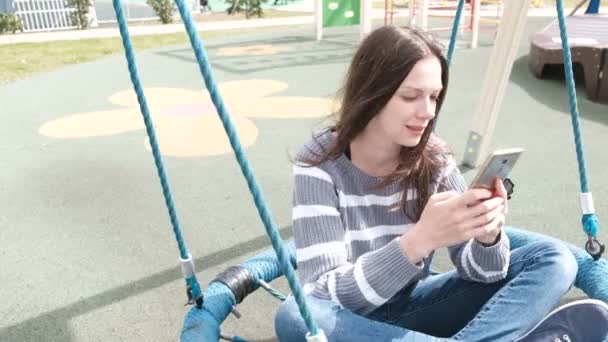 Όμορφη μελαχρινή γυναίκα ταλαντεύεται στο άδειο webbed swing, loking στο social media στο κινητό τηλέφωνο. — Αρχείο Βίντεο