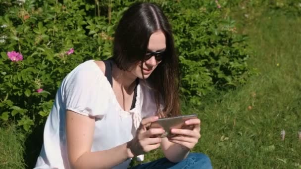 Mulher está jogando um jogo no telefone móvel sentado no parque em dia ensolarado . — Vídeo de Stock