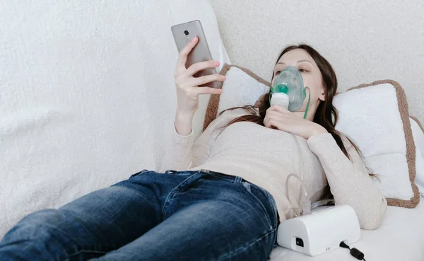 Используйте для лечения ингалятор и распылитель. Молодая женщина, вдыхающая через маску ингалятора, лежит на диване и болтает в мобильном телефоне. Вид спереди . — стоковое фото