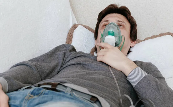 Use nebulizador e inhalador para el tratamiento. Hombre joven inhalando a través de la máscara del inhalador acostado en el sofá. Vista frontal de primer plano . — Foto de Stock