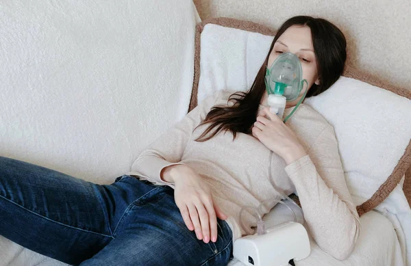 Use nebulizador e inhalador para el tratamiento. Mujer joven inhalando a través de la máscara del inhalador acostada en el sofá. Vista frontal . — Foto de Stock