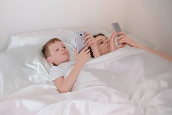 Mamá e hijo se despiertan y navegan por Internet en sus teléfonos móviles. Vista lateral . — Foto de Stock