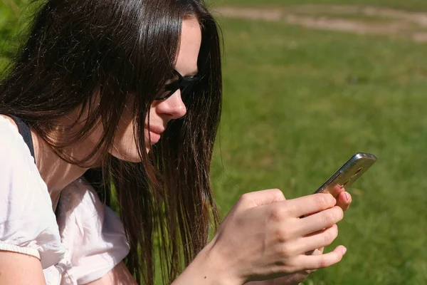 La mujer está escribiendo un mensaje en el teléfono móvil sentado en el parque en el día soleado. Vista lateral . — Foto de Stock