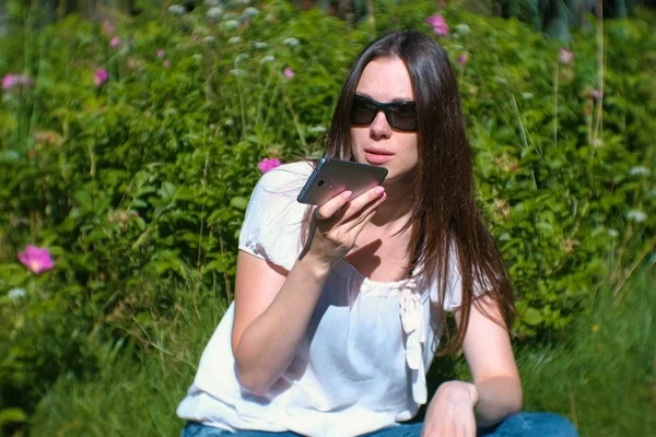 Mulher morena bonita grava uma mensagem de voz em seu telefone celular enquanto está sentada no parque em um dia ensolarado . — Fotografia de Stock