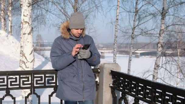 Hombre de chaqueta azul con capucha de piel usando su celular para la web en un parque de invierno . — Vídeos de Stock