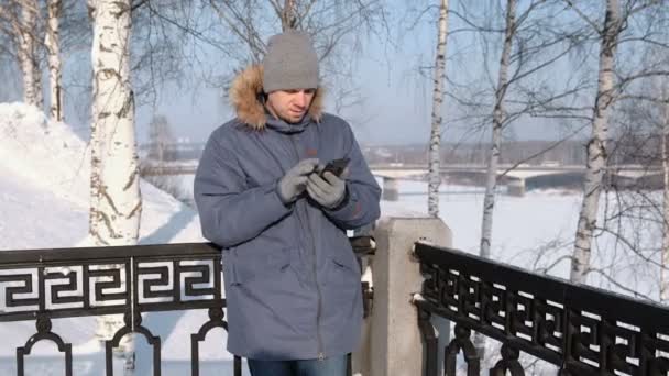 Hombre de chaqueta azul con capucha de piel limpia la pantalla del teléfono con la mano en guantes en un parque de invierno . — Vídeos de Stock