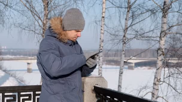 Hombre de chaqueta azul con capucha de piel quita un guante usando su teléfono celular para la web en invierno Park . — Vídeos de Stock