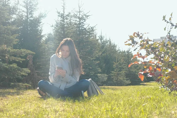 젊은 아름 다운 갈색 머리 여자 봄 공원에서 잔디에 앉아서 휴대 전화에서 메시지 유형. — 스톡 사진