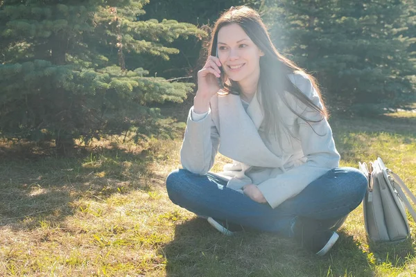 Retrato de la joven feliz hermosa mujer sentada en el parque de primavera en la hierba y hablando en un teléfono móvil . — Foto de Stock