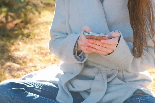 Mulher morena bonita jovem senta-se na grama no parque da primavera e digita uma mensagem no telefone celular. Mãos fechadas . — Fotografia de Stock