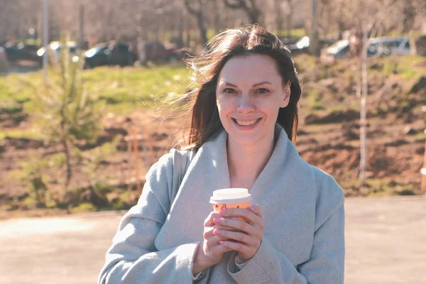 Retrato de una joven morena en abrigo pasea por el parque de la ciudad y bebe café. Primavera . — Foto de Stock