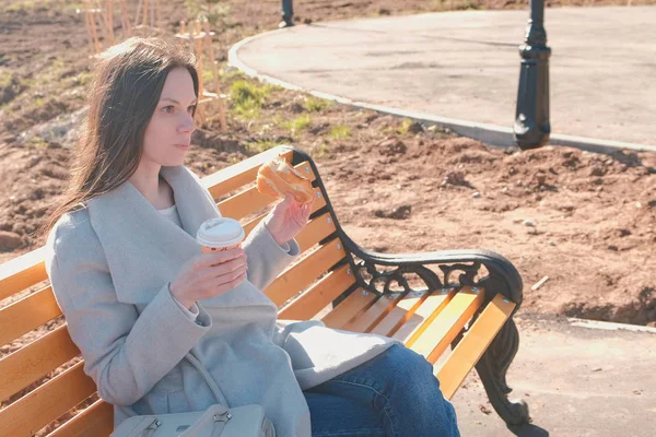Молодая брюнетка в пальто сидит на скамейке в городском парке, ест булочку с сахаром и пьет кофе. Весна . — стоковое фото