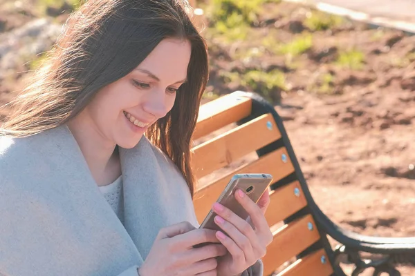 Portrett av lykkelig brunette som leser og skriver en beskjed på telefonen sittende på benken i parken . – stockfoto