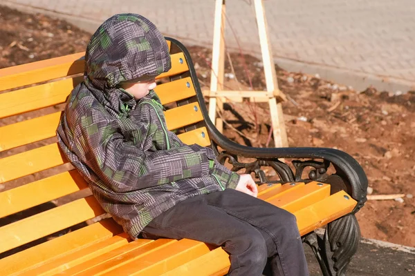 Rapaz senta-se num banco no Parque e coça o banco com uma pedra . — Fotografia de Stock