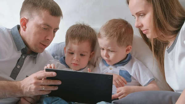 Family with tablet. Mom, dad and two sons twins toddlers looking cartoon at tablet lying on the bed.