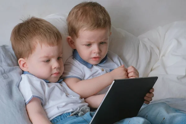 Дети с планшетом. Два мальчика близнецы малыши смотрят мультфильм на планшет, лежащий на кровати . — стоковое фото