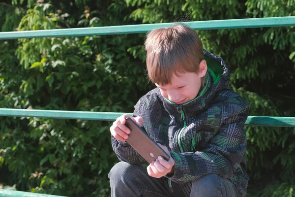 A fiú játszik egy játék a mobiltelefonon a parkban ülve. — Stock Fotó